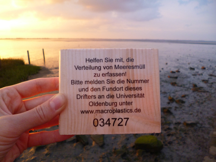 Holztafel mit der Aufschrift: Helfen sie mit die Verteilung von Meeresmüll zu erfassen! Bitte melden sie die Nummer und den Fundort dieses Drifters an die Universität Oldenburg unter: www. macroplastics.de Nummer: 03472