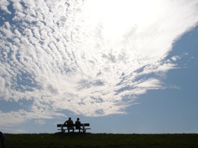 two people sit next to each other on a bench under a sunny and cloudy sky